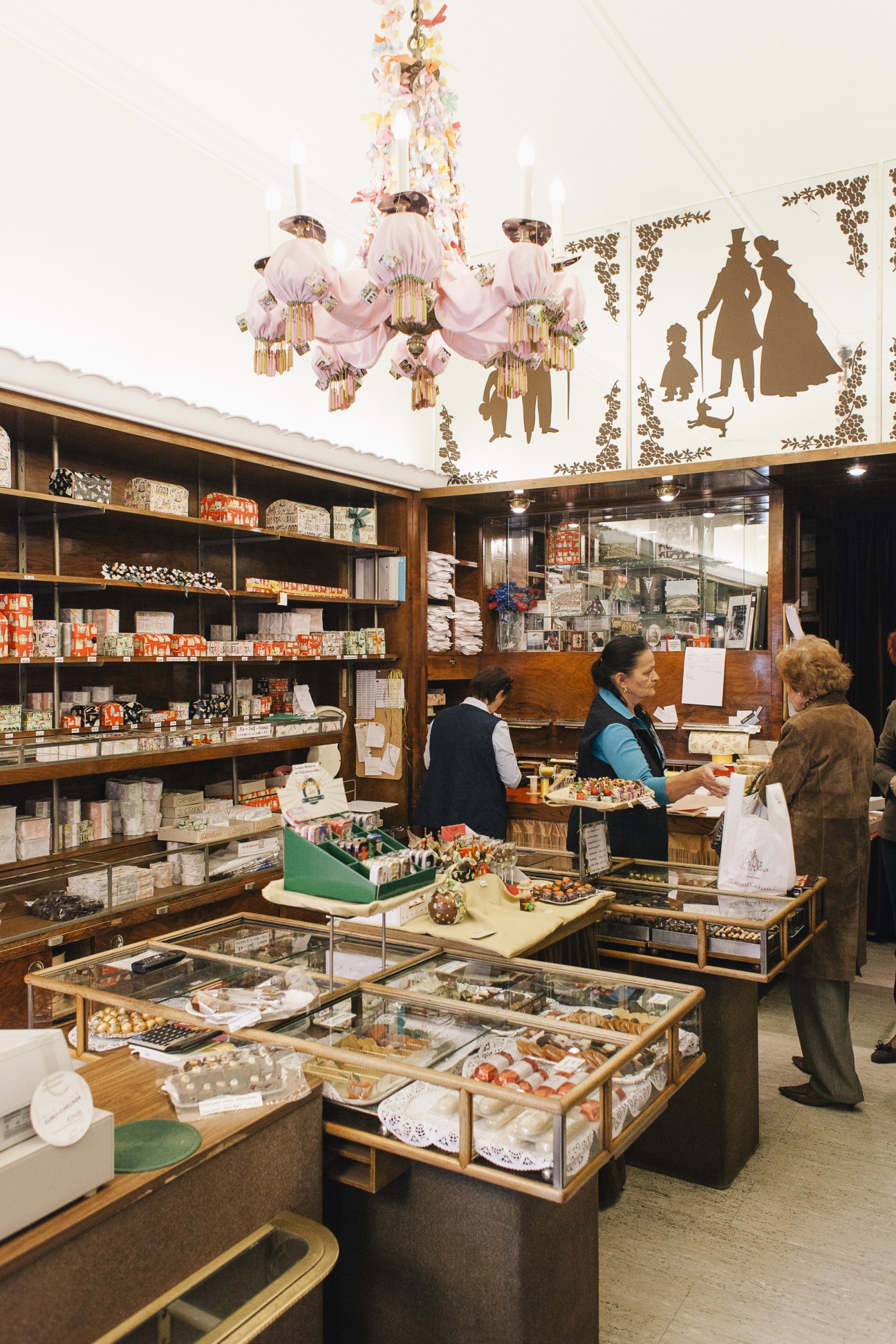 Interior of Altmann & Kühne confectionery shop with wooden display cases, pink chandelier, and customers browsing handmade chocolates.