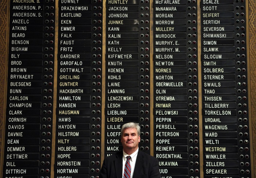 Man in suit and striped tie standing in front of a large wall display of names with checkboxes.