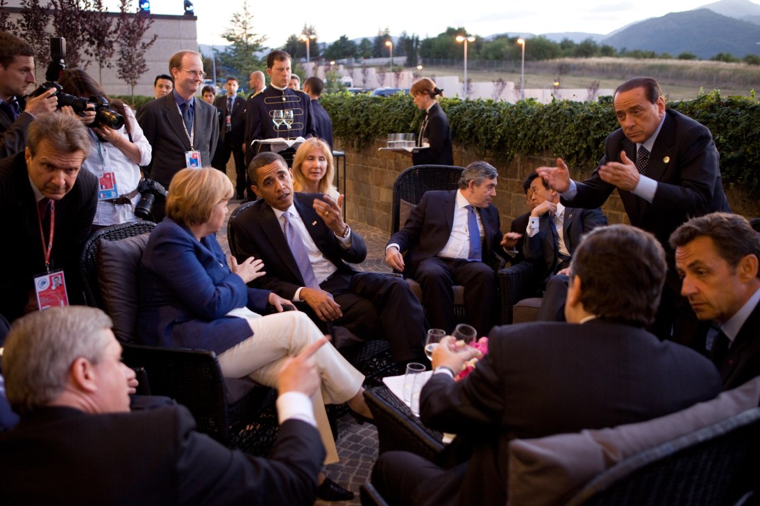 International leaders and diplomats at an outdoor gathering with mountains in background, including Angela Merkel and Barack Obama.