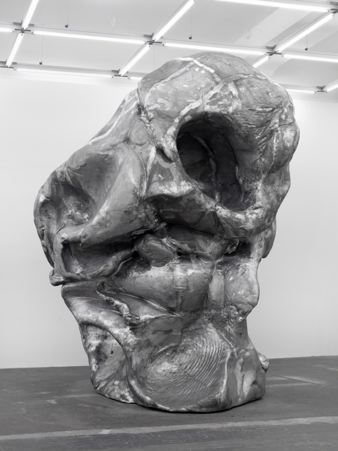 Large abstract metal sculpture with hollow cavities displayed in a white gallery space with fluorescent ceiling lights.