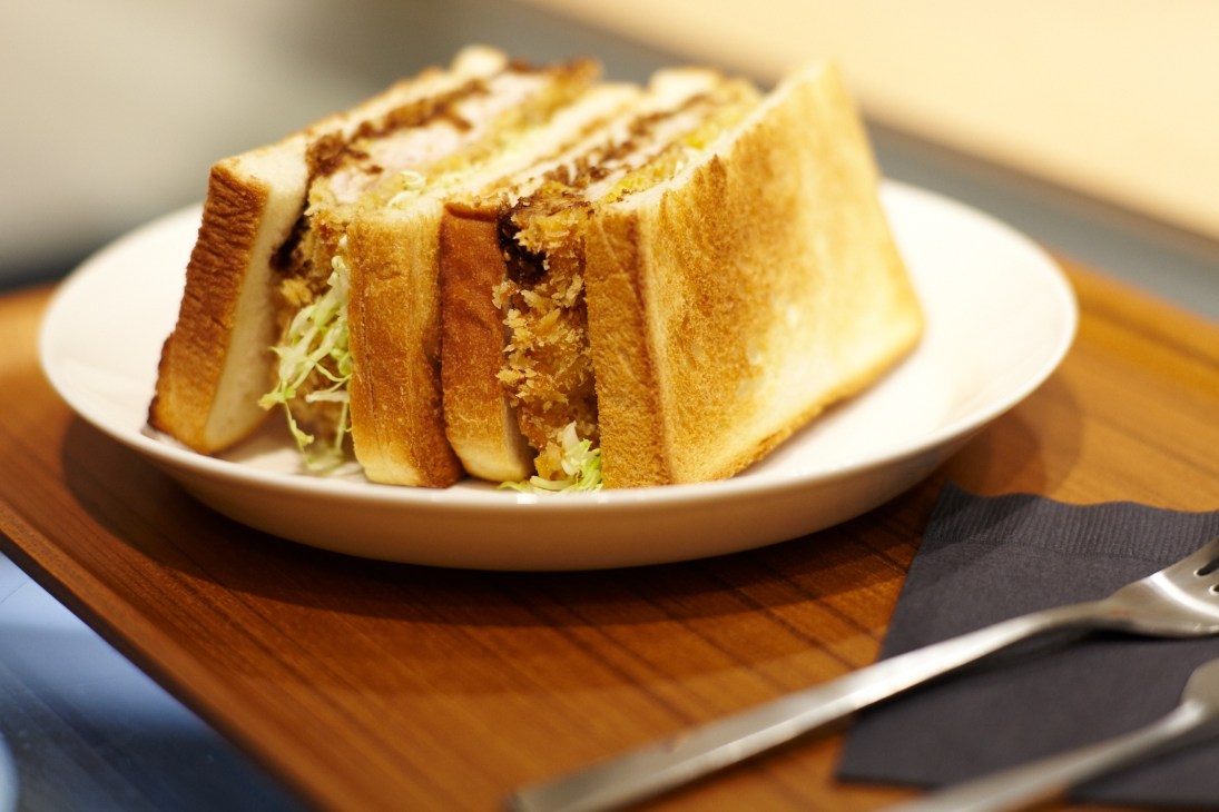 Toasted sandwich with pork cutlet and greens on a white plate with wooden board and cutlery.
