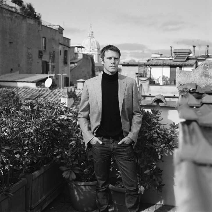 Prince Emanuele Filiberto di Savoia stands on a rooftop terrace with potted plants, overlooking a European cityscape with a domed building in the background.