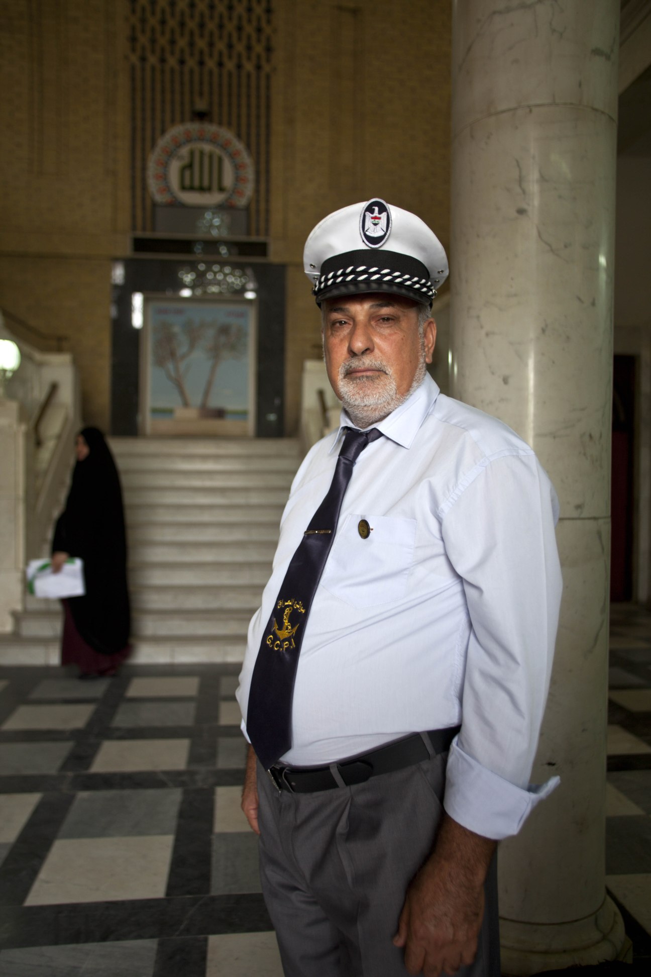 Hussein Ali, a guard of the State Company for Iraqi Ports, wearing a white uniform and cap in an institutional corridor.
