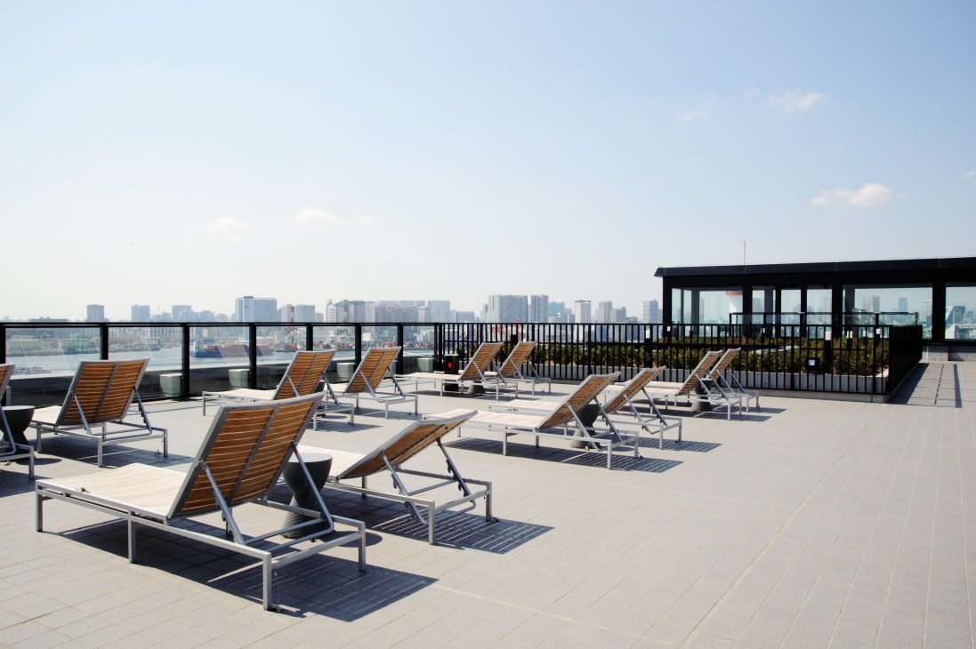 Sun loungers with wooden seats on a rooftop terrace overlooking Tokyo Bay and the city skyline.