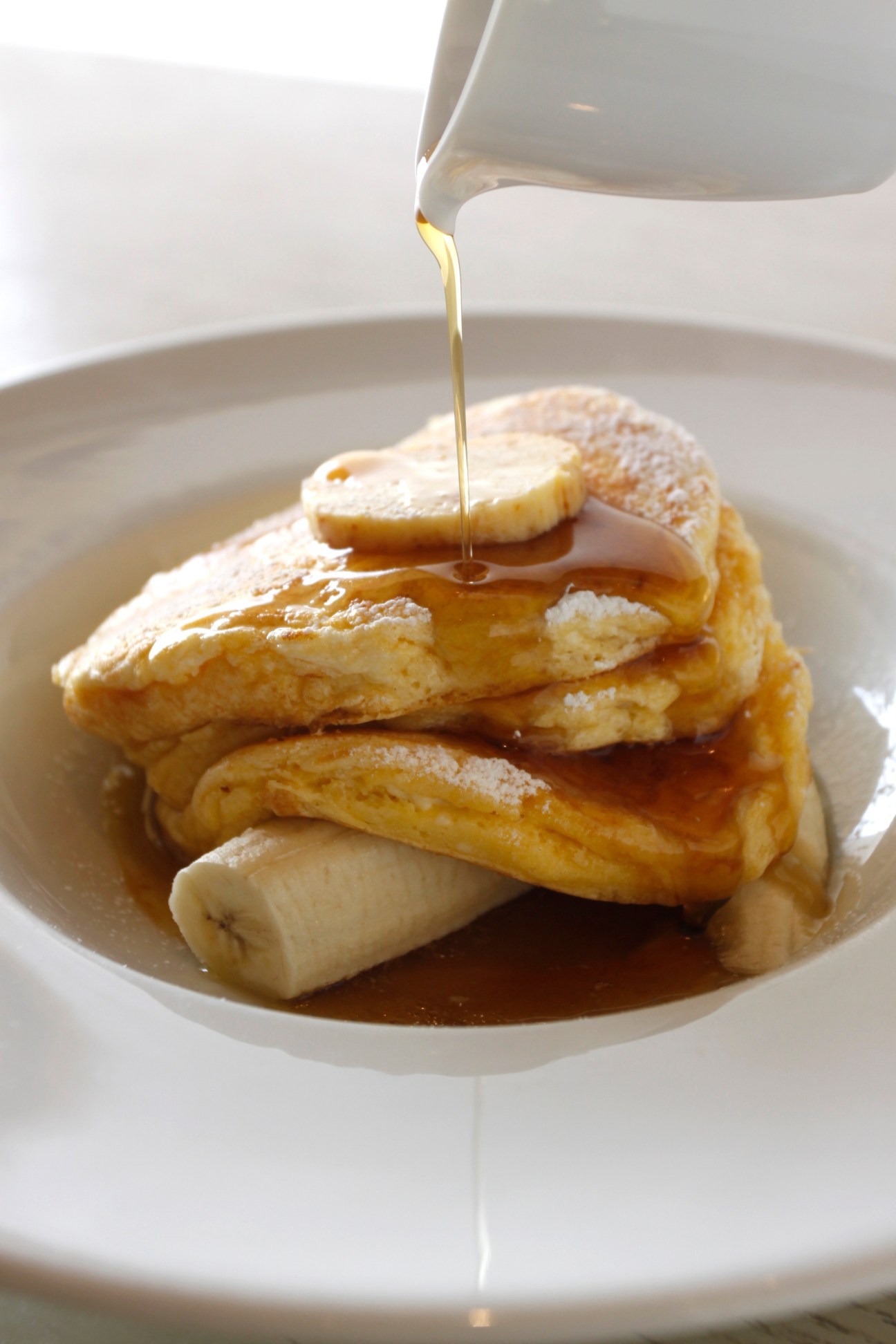 Syrup being drizzled onto a stack of fluffy pancakes with a sliced banana at Bills restaurant.