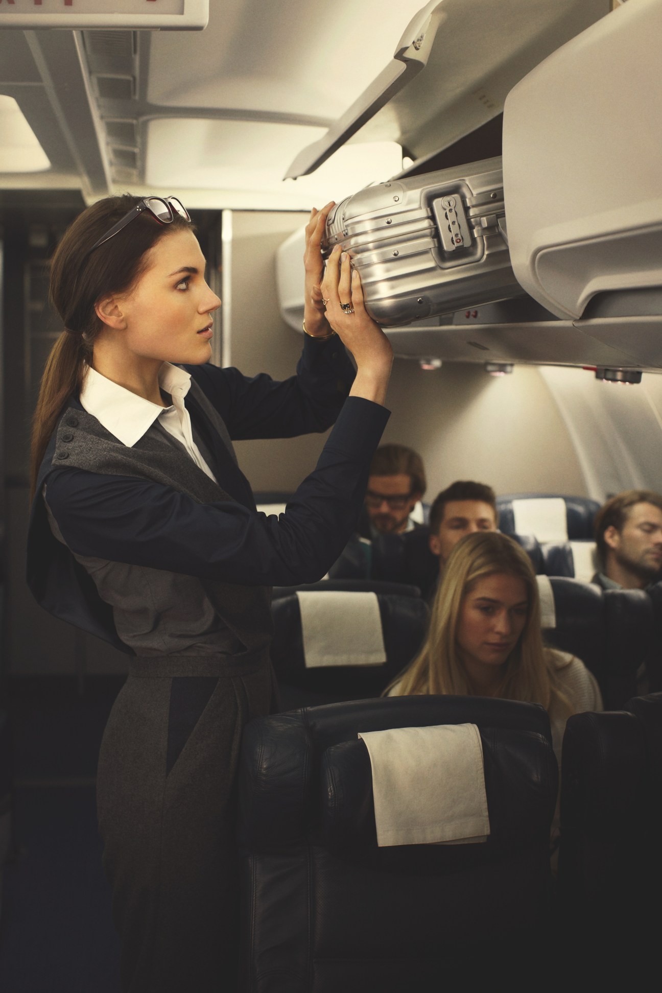 Woman in airplane aisle placing Rimowa suitcase in overhead bin, wearing Oliver Peoples glasses and 1205 jumpsuit.