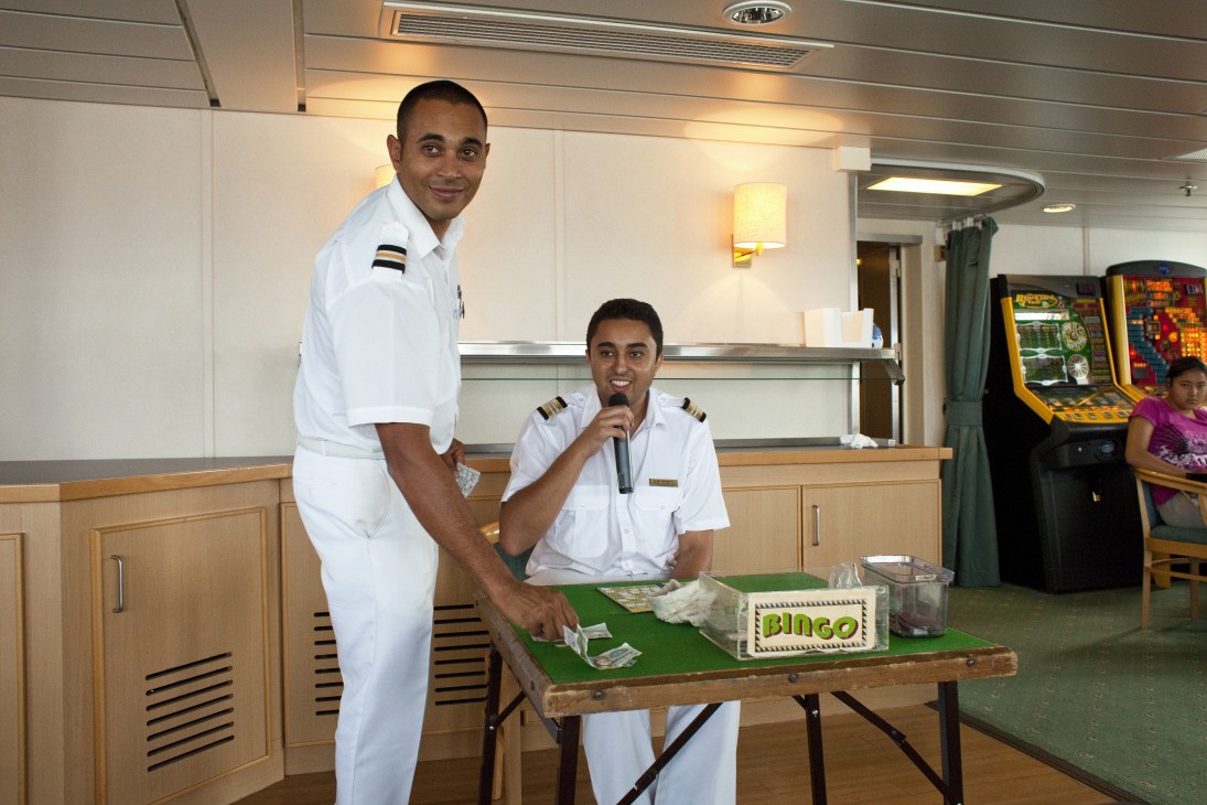 Two ship officers in white uniforms run a bingo game in the main lounge of the RMS St Helena.
