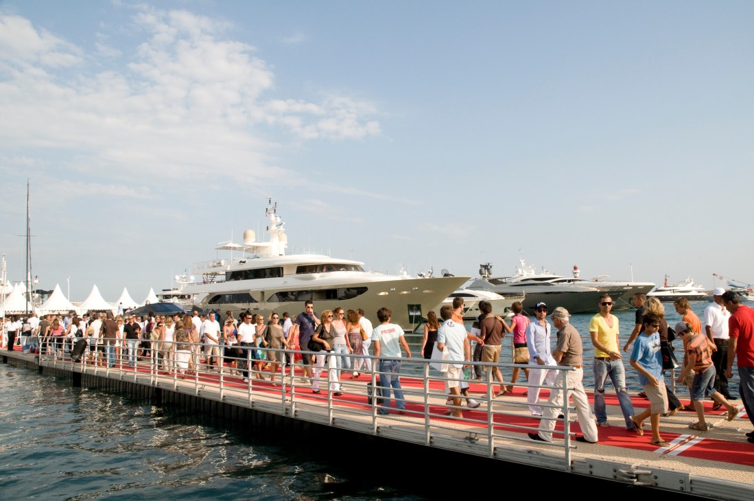 Visitors queue on a red-carpeted bridge at a yacht show with luxury motor yachts moored behind them.