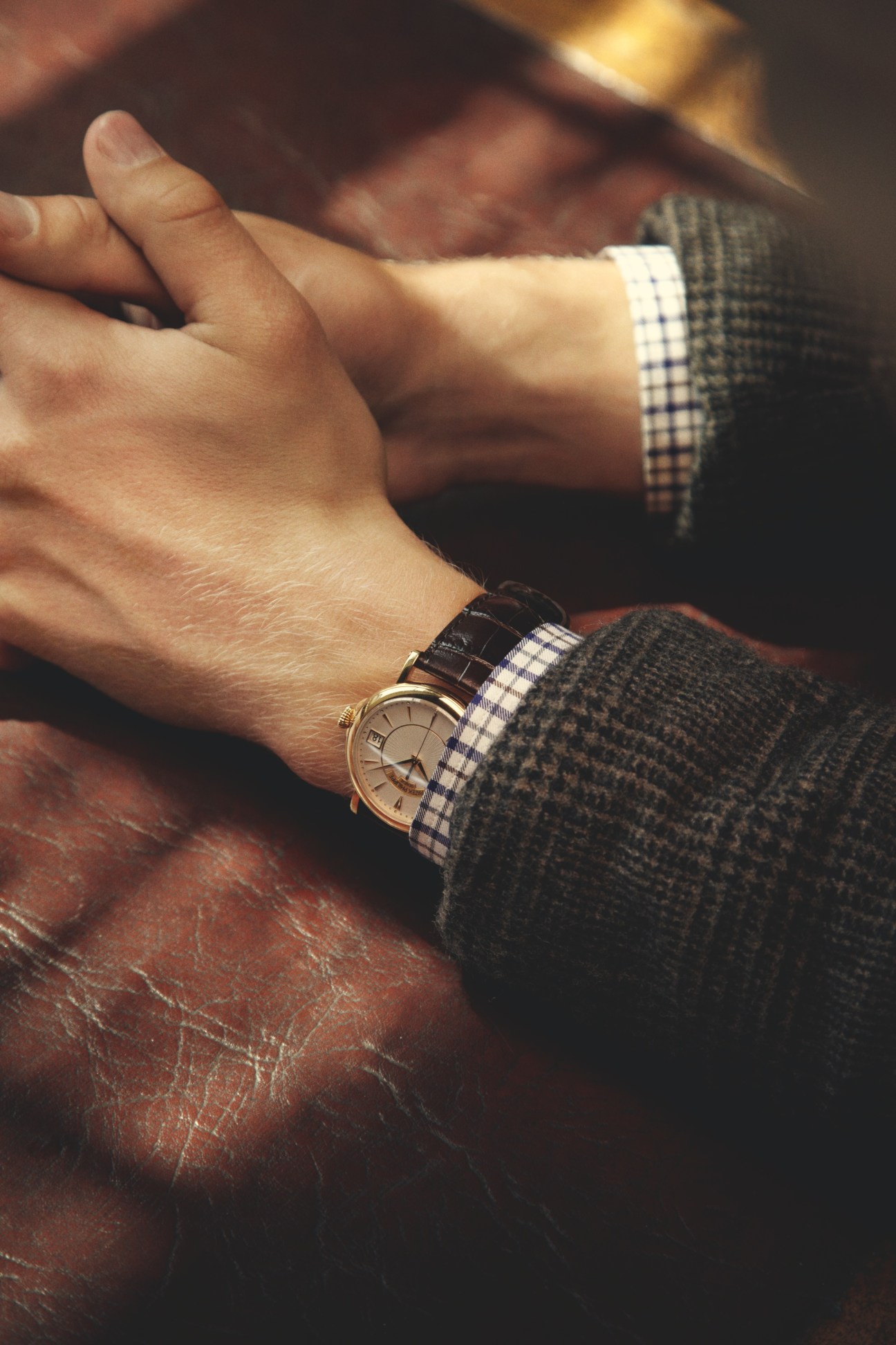 Close-up of a Patek Philippe watch on a wrist wearing a checkered shirt and dark jacket.