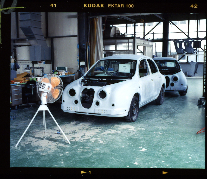 White car body in assembly stage at Mitsuoka factory with fan and industrial workshop visible.
