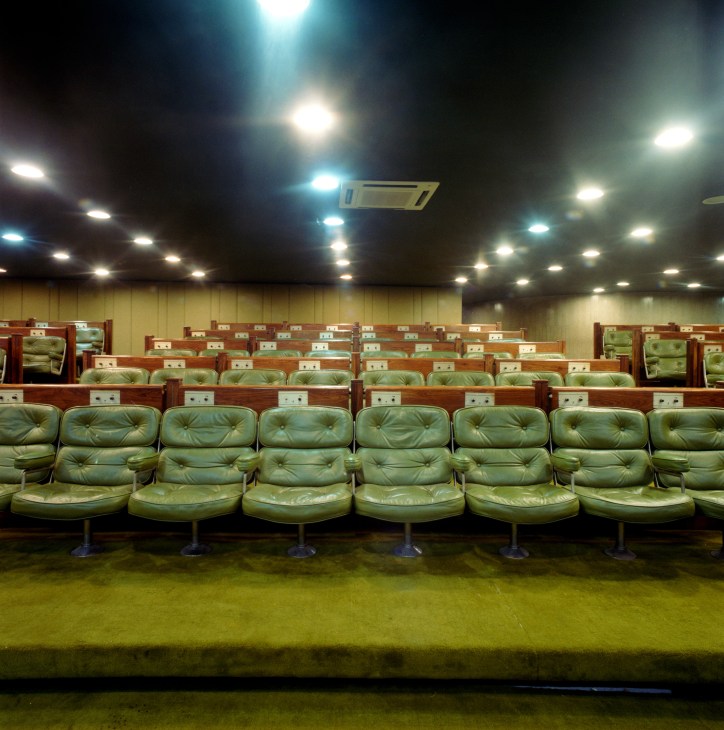 Press conference room with rows of green leather seats and wooden partitions under bright ceiling lights.