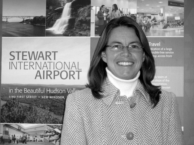 Portrait of Dianne Ehler, General Manager of Stewart International Airport, smiling at camera in front of promotional materials.