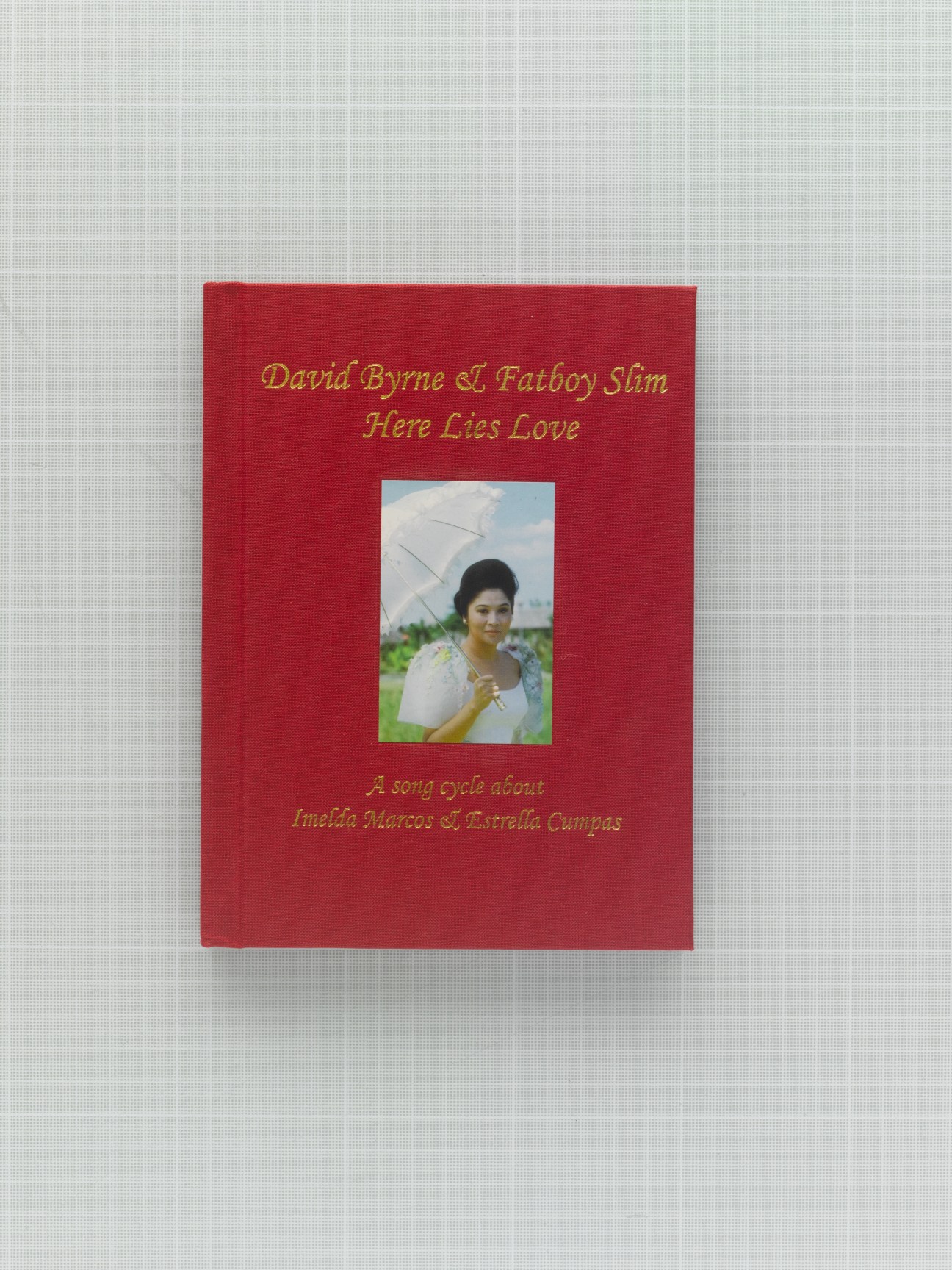 Red book cover for David Byrne & Fatboy Slim's 'Here Lies Love' album featuring gold text and a photograph of a woman with an umbrella.