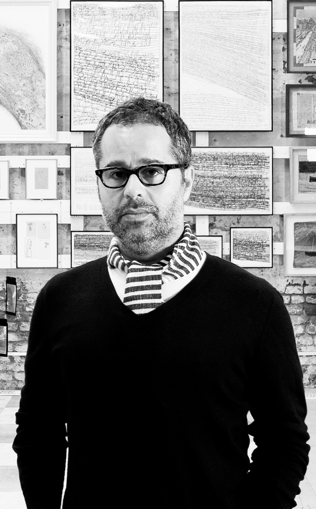 Man with glasses and striped scarf standing in front of wall displaying framed architectural drawings and sketches