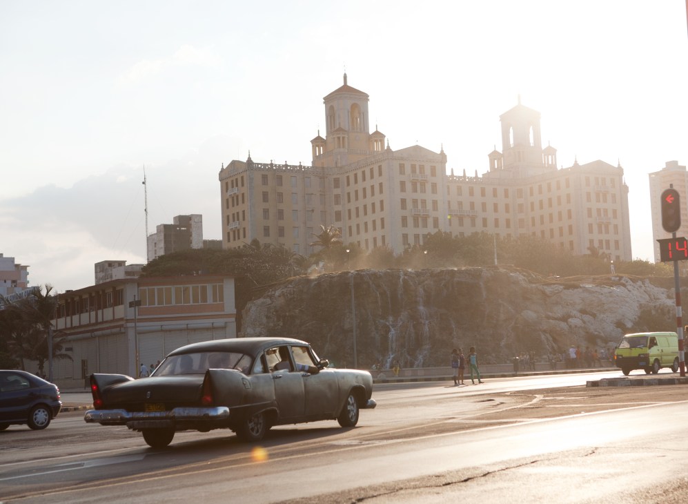 Classic American car parked on Havana street with Hotel Nacional and colonial buildings in background.