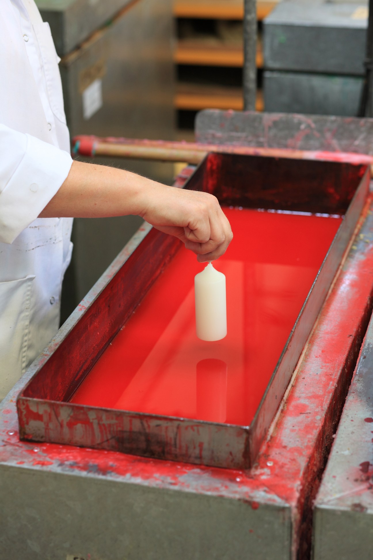 Worker's hand dipping a white pillar candle into a red dye bath at Cire Trudon factory.