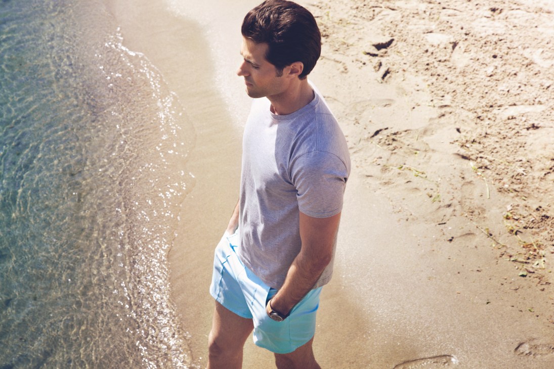 Man in gray t-shirt and turquoise swim trunks standing on a sandy beach by the water.