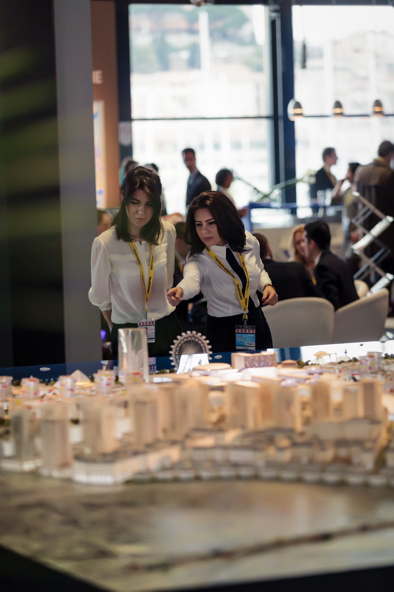 Two women in white shirts and lanyards examine a large architectural model at a trade show booth.