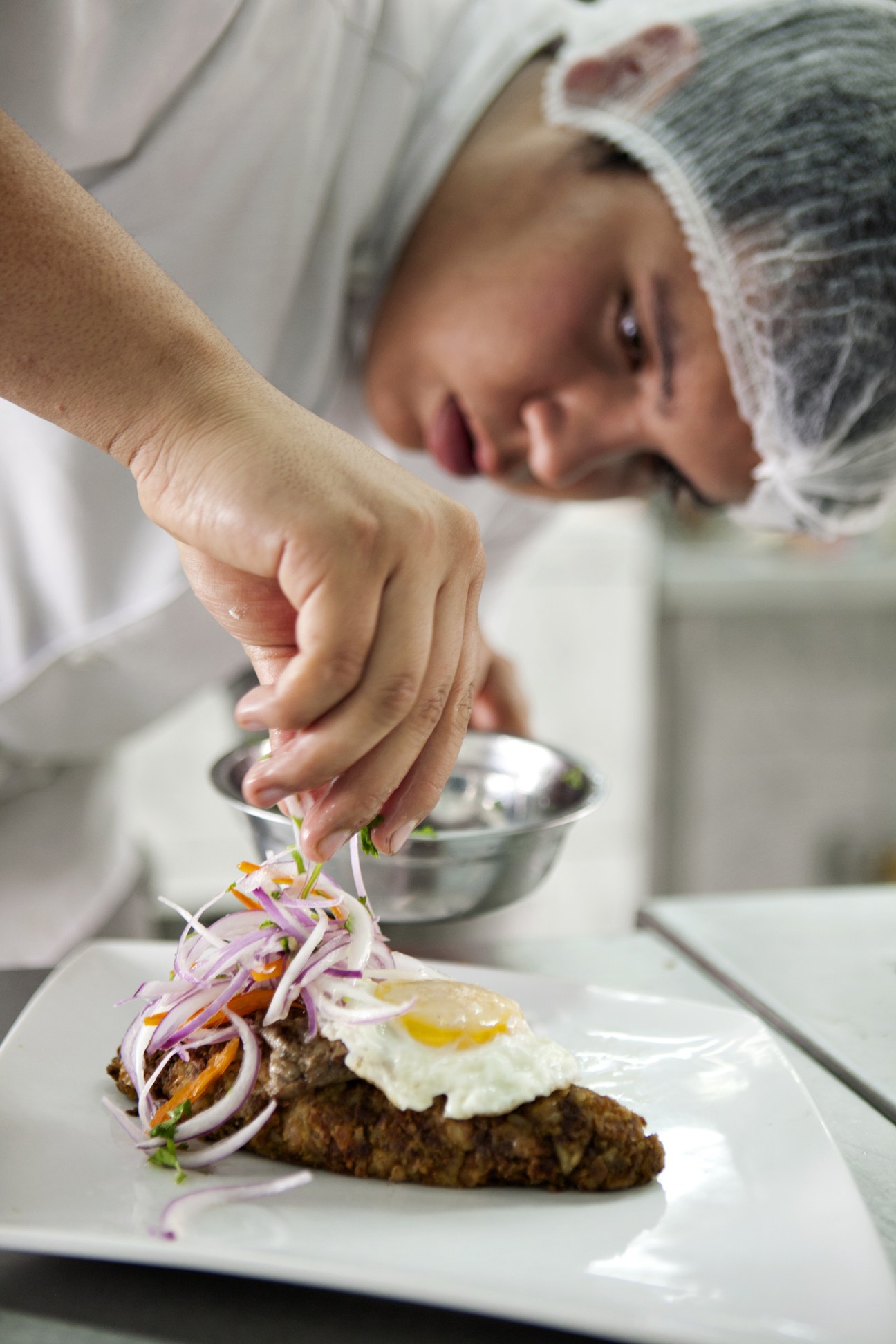 A chef in white hat and coat garnishes a plate of tacu-tacu with purple onion slices.