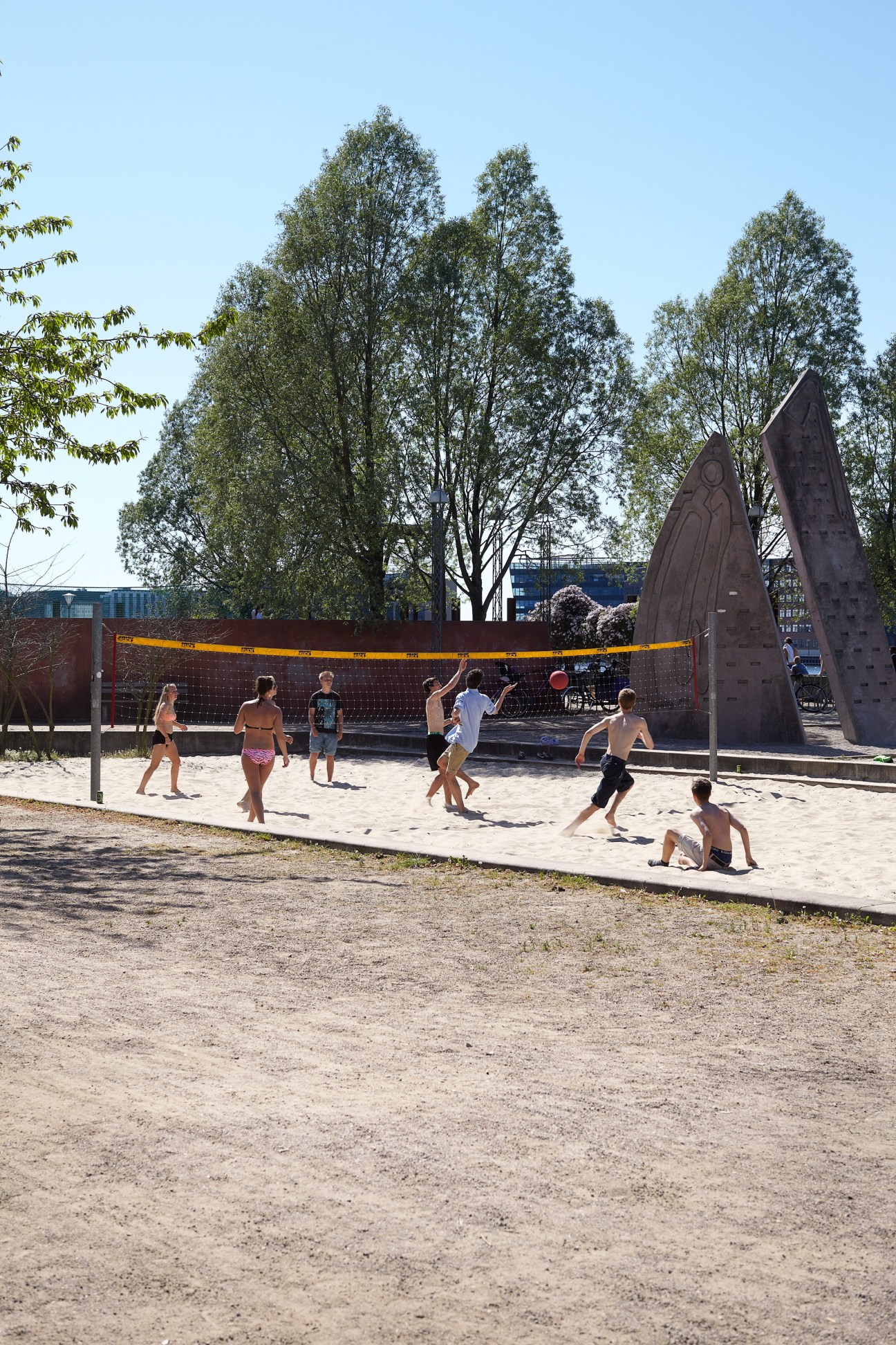 Children playing beach volleyball on sand with tall trees and modern sculptures in background.