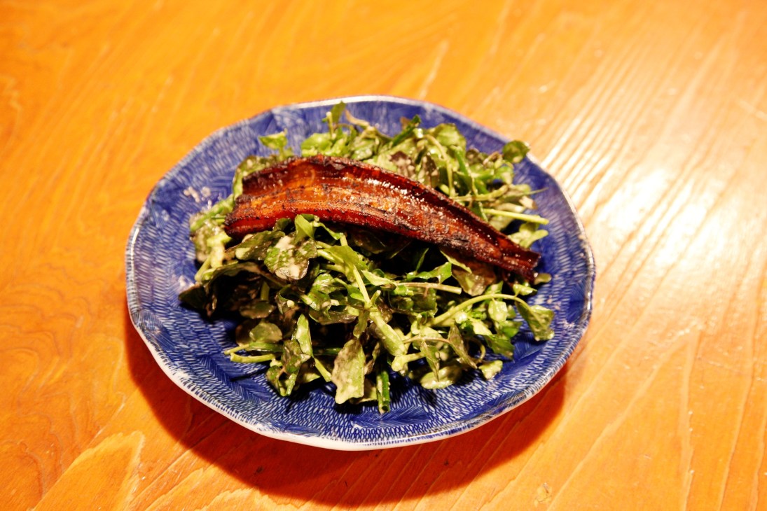 Grilled sea eel on a bed of watercress in a blue patterned bowl on a wooden surface.