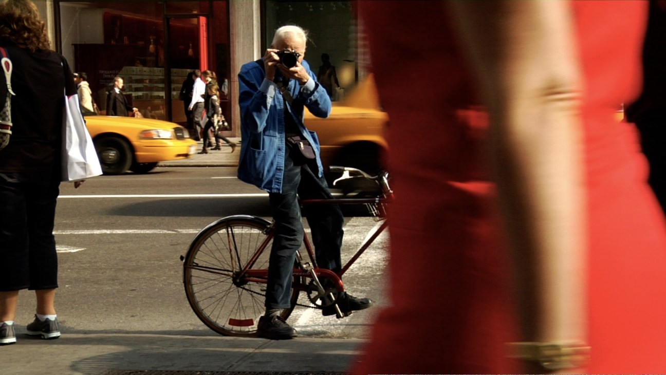 Elderly man on bicycle with camera on New York City street with yellow taxi