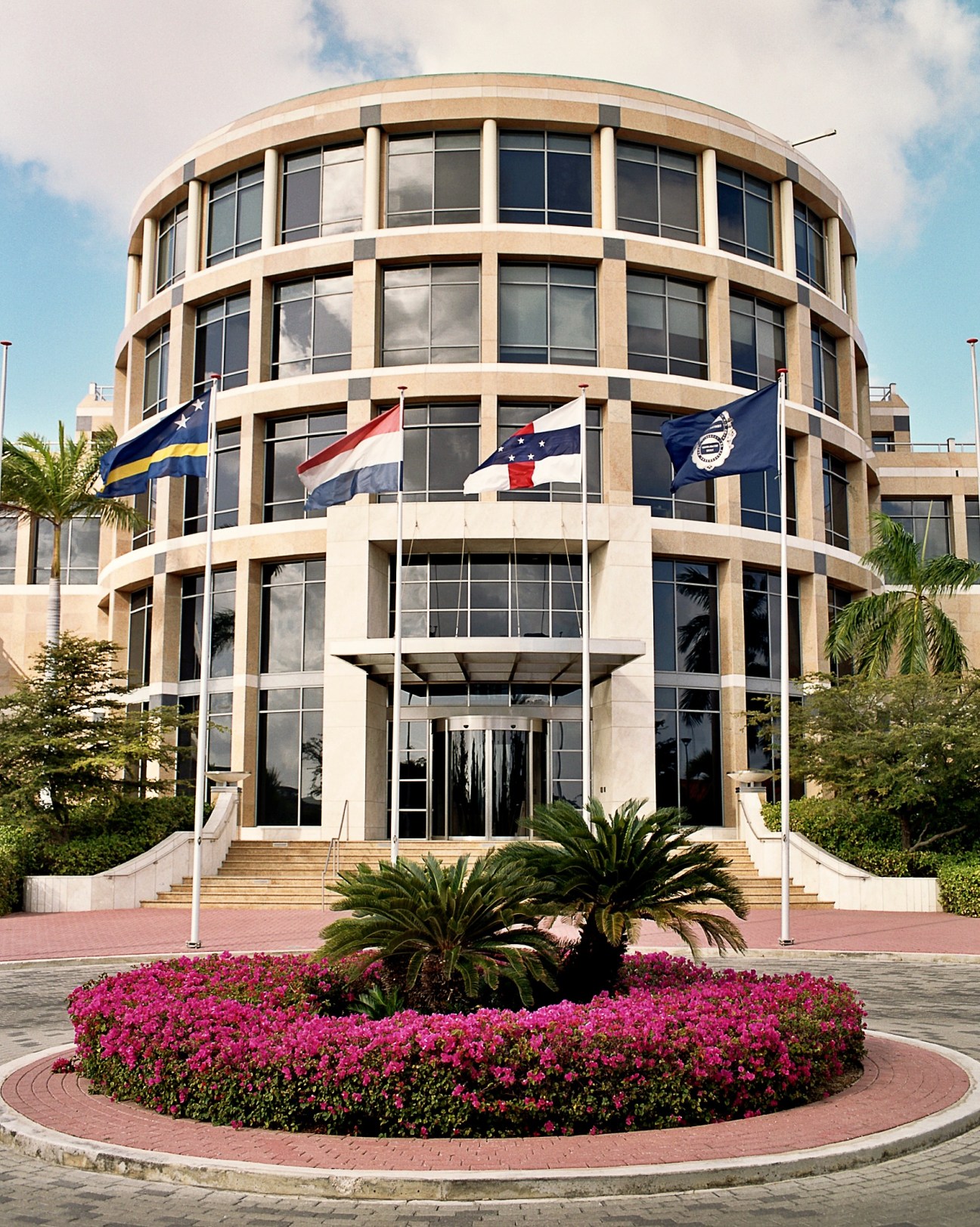 Cylindrical modern building with flags of Curaçao, Netherlands, Netherlands Antilles, and central bank crest flying from its facade.