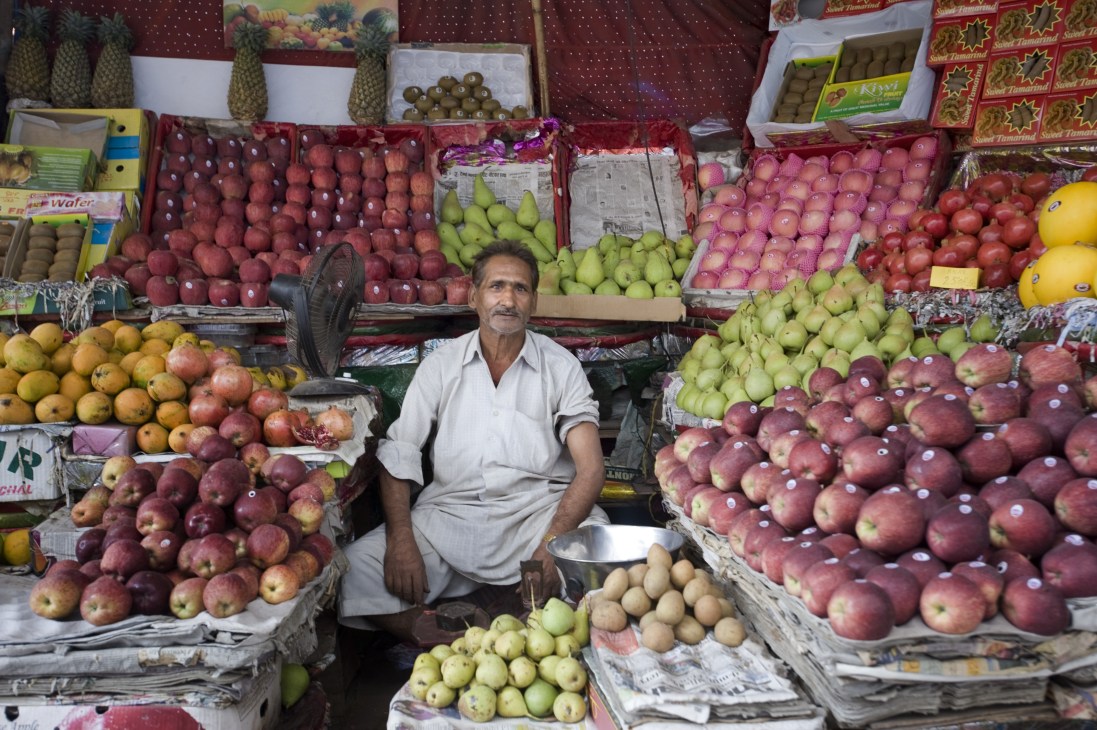 Fruit vendor surrounded by colorful displays of apples, pears, and other fresh produce at his Old Delhi market stall.