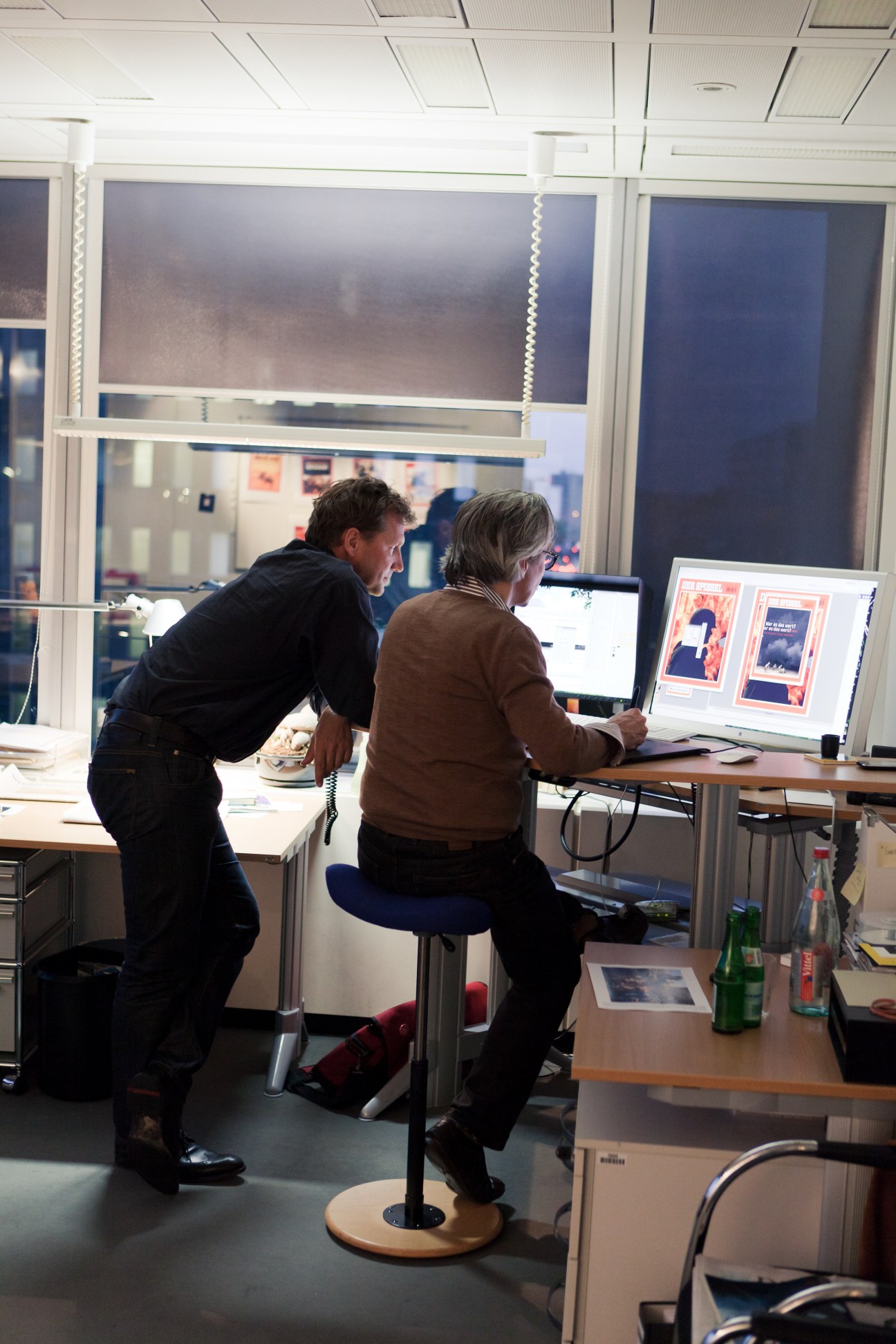 Two men work at a desk reviewing magazine cover designs on a computer screen.