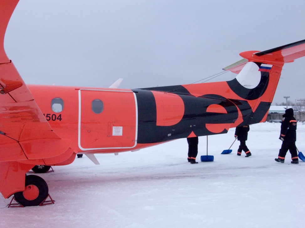 Ground crew prepare a bright orange and black Pilatus PC-12 on a snowy runway at Bykovo Airport in winter.