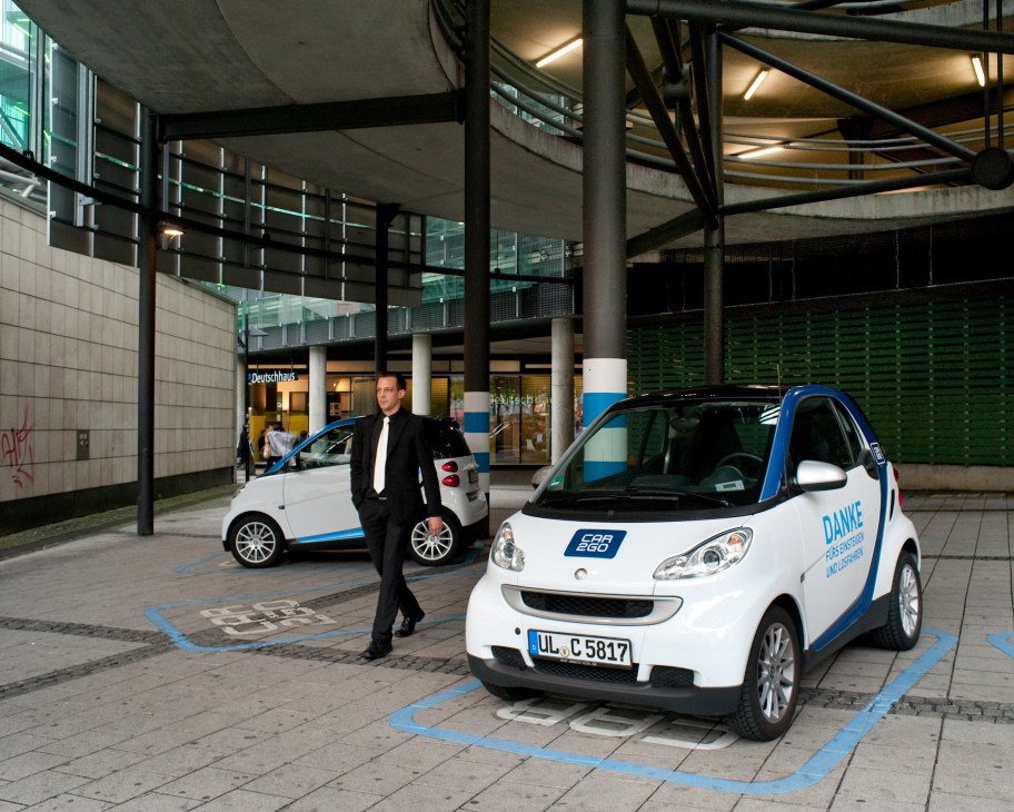 Man in black blazer stands between two white and blue Smart cars parked in a modern urban courtyard with concrete pillars.