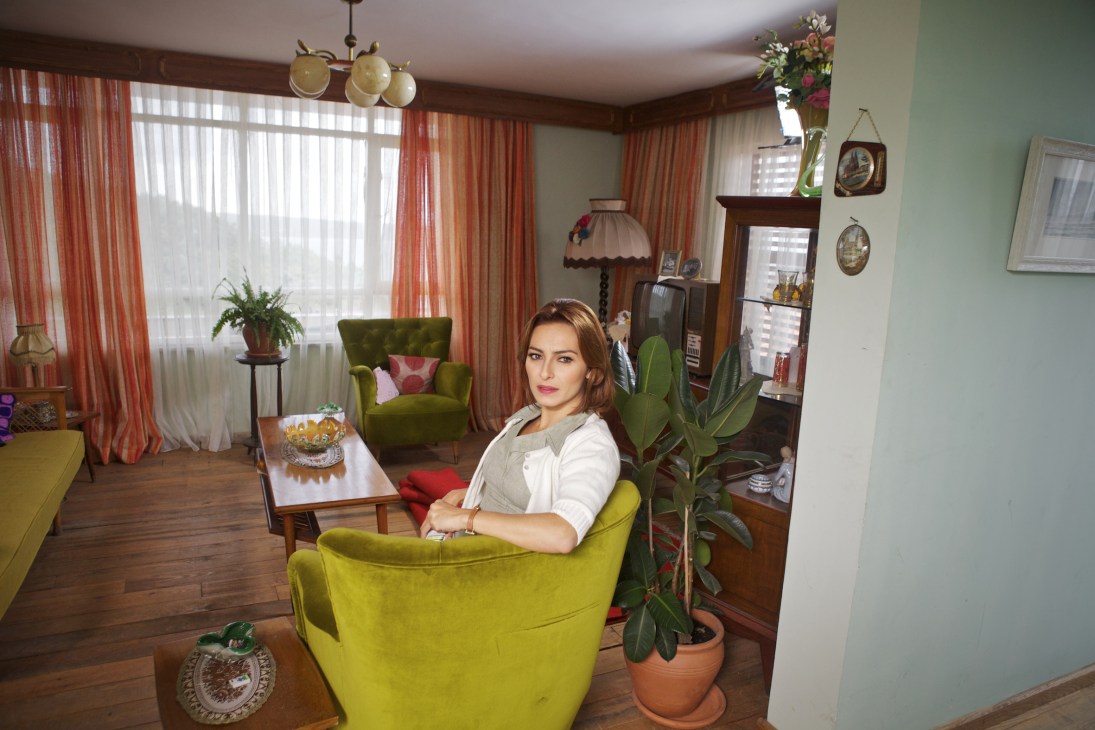 Actress Ayça Bingöl sits in a yellow armchair in a stylishly decorated living room set.