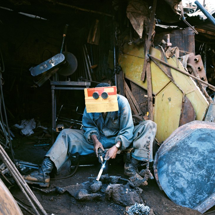 Welder at Medeber market in Asmara wearing protective goggles and cardboard shield while working with metal scraps.
