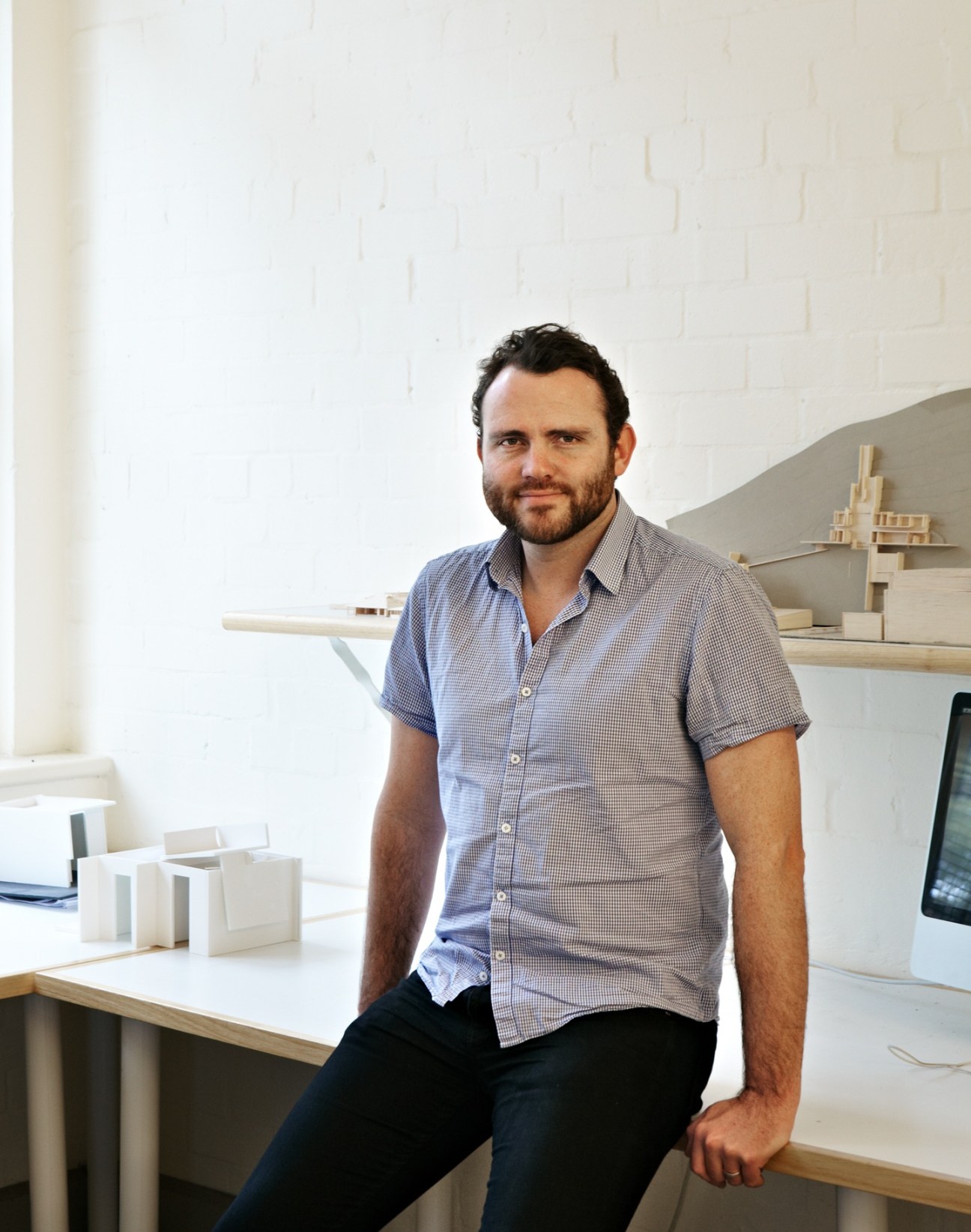 Andrew Burns, architect, posing in his minimalist office with architectural models on shelves behind him.