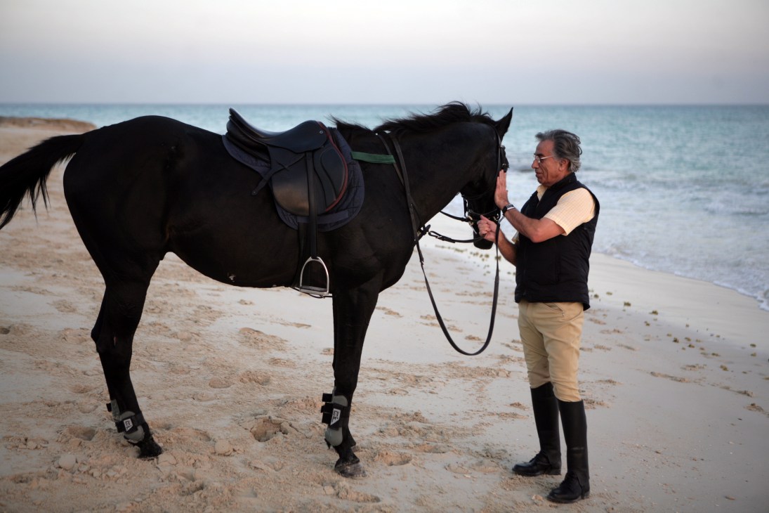 Man in equestrian riding gear prepares a black horse for photos on Kish beach at sunset.