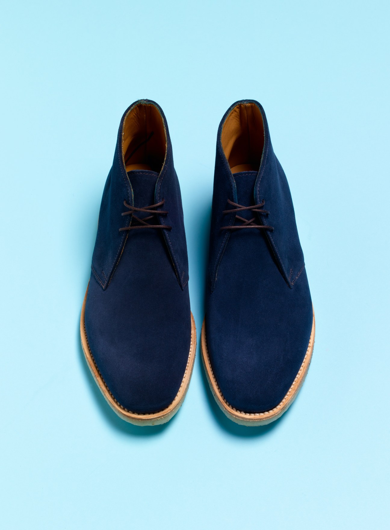 Pair of navy blue suede desert boots with brown leather laces and tan crepe sole against light blue background.