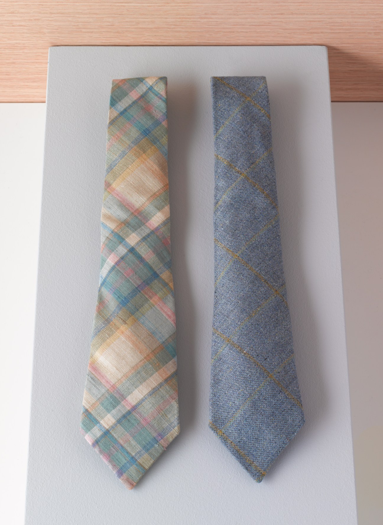 Two neckties displayed on white paper—one with a multicolored plaid pattern and one solid blue with geometric design.
