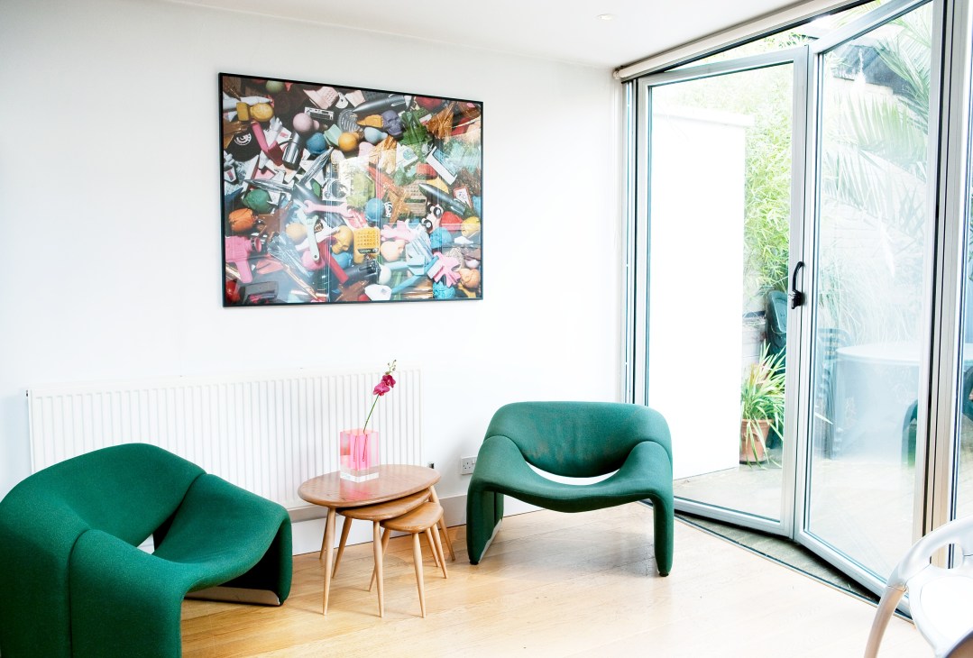 Modern interior space with green sculptural chairs, wooden side table, and colorful collage artwork on white wall.