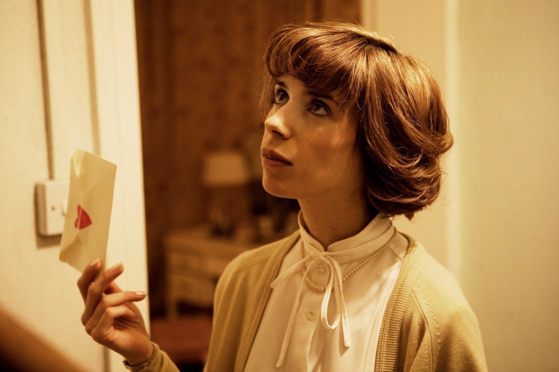 A woman with short brown hair wearing a light yellow cardigan holds a postcard while looking to the side in an interior setting.