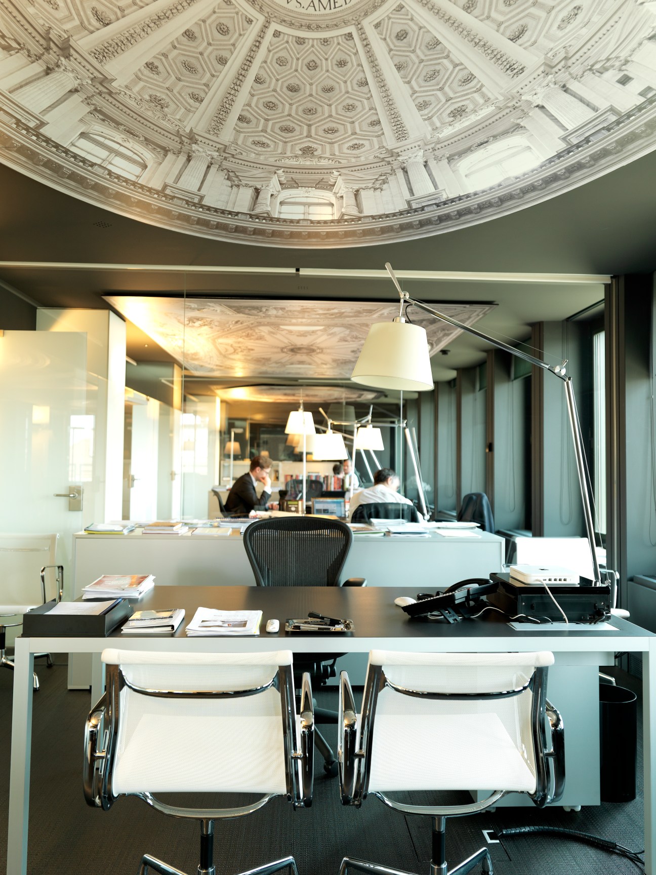 Office workspace with ornate ceiling mural, white desks, black chairs, and arched window overlooking the city.