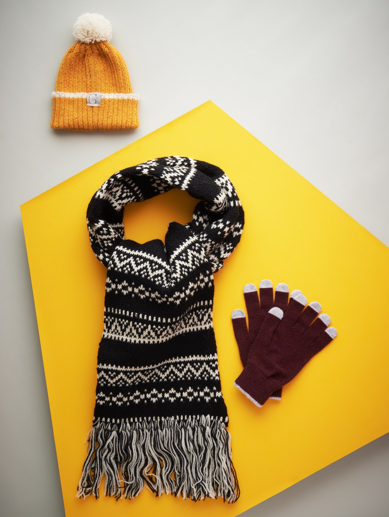 Orange beanie with cream pom-pom, black and white patterned scarf, and burgundy gloves with touch-screen compatible fingertips on yellow background.