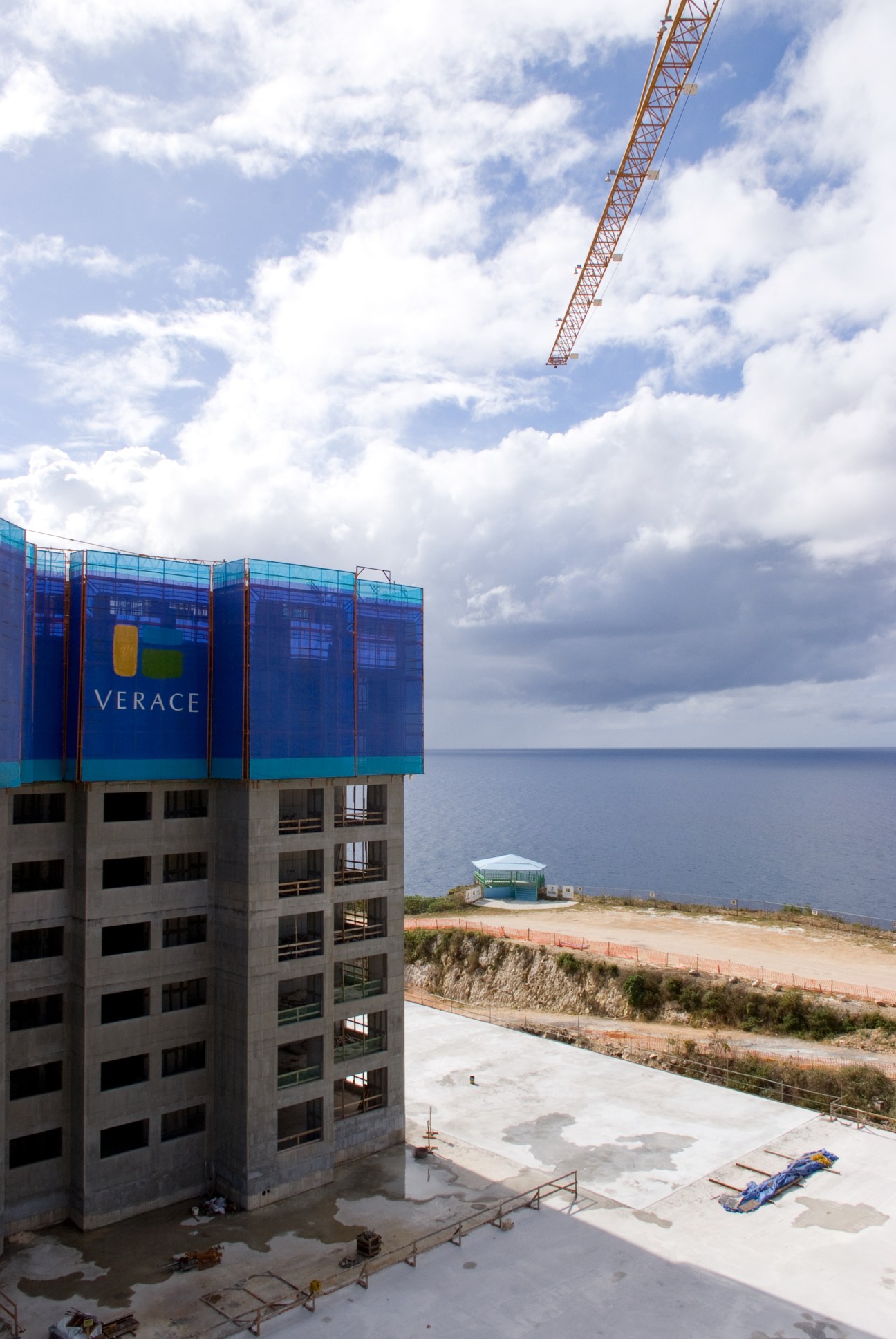Construction crane above an unfinished residential tower with blue signage reading 'VERACE' overlooking ocean and beach.