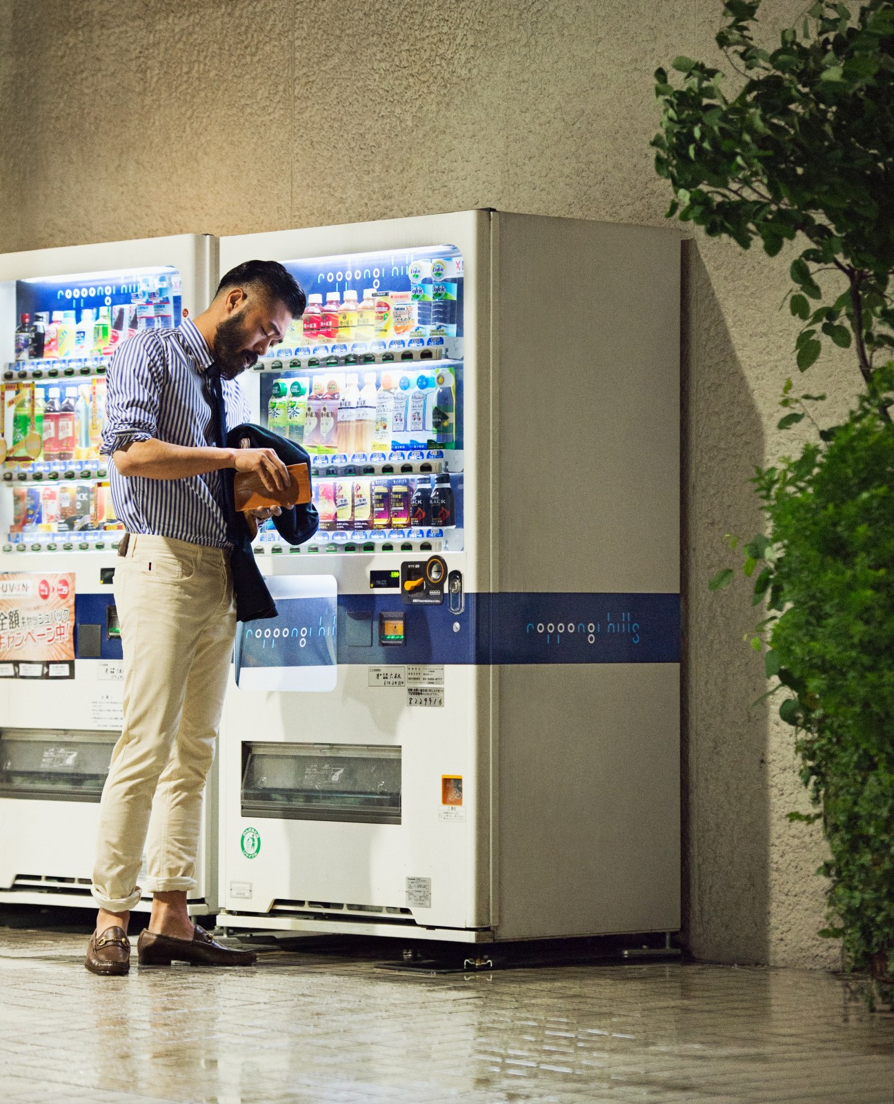 Man in striped shirt and cream trousers using a Japanese vending machine stocked with colorful beverages.