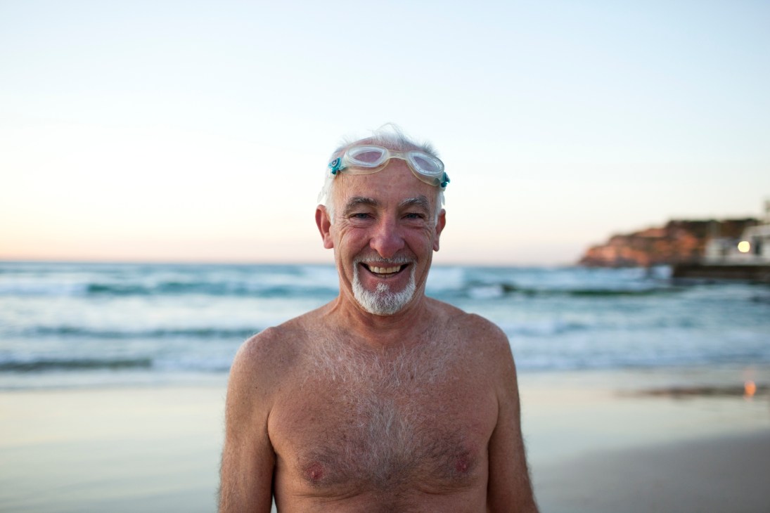 Older man with white beard wearing swimming goggles on his head, smiling at Bondi Beach at sunrise with waves behind him.