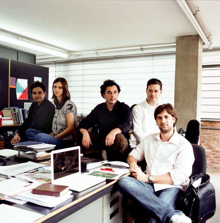 Five professionals posing in a modern office with concrete column and shelving, representing Arquitetos Associados.