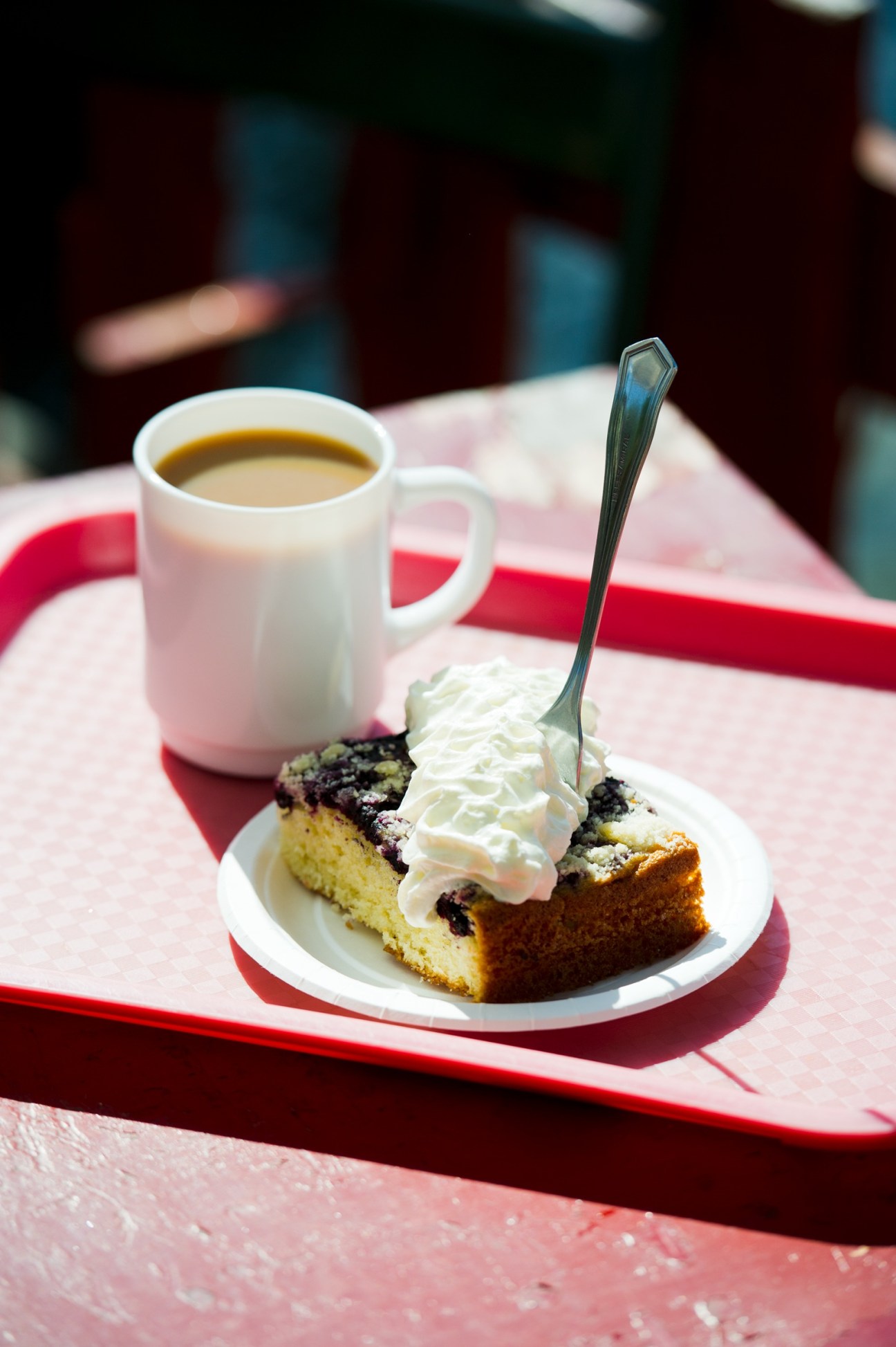 A slice of berry cake with whipped cream served on a white plate next to a white mug of coffee on a red tray.