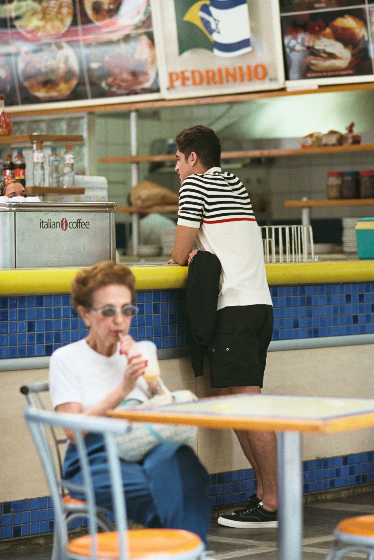 Man in striped polo shirt and shorts at a café counter with Italian Coffee signage visible.
