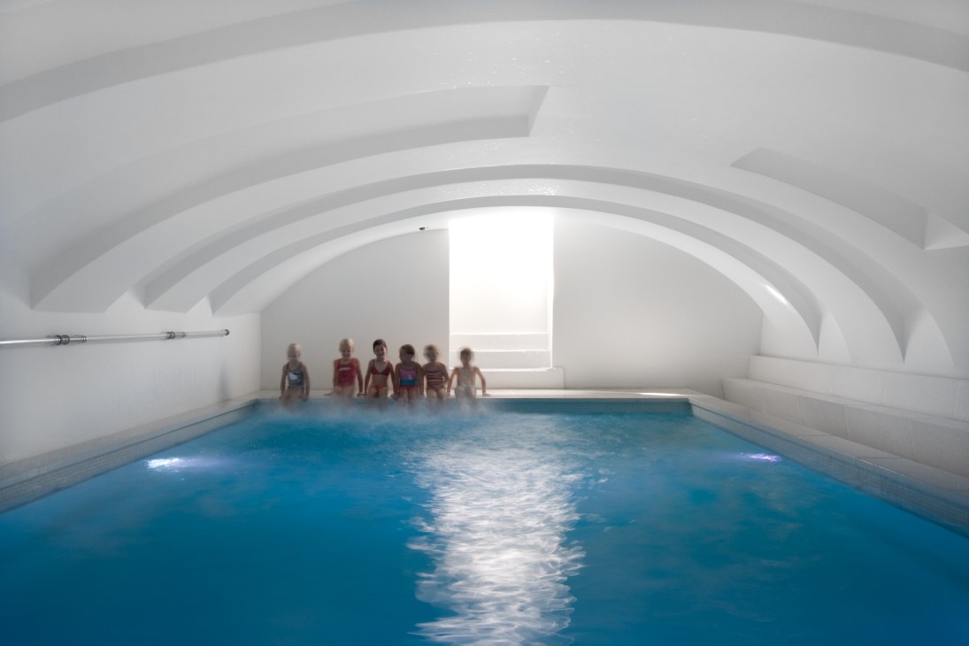 Children stand at the edge of a blue swimming pool inside Hotel Zenden's white vaulted medieval chamber.