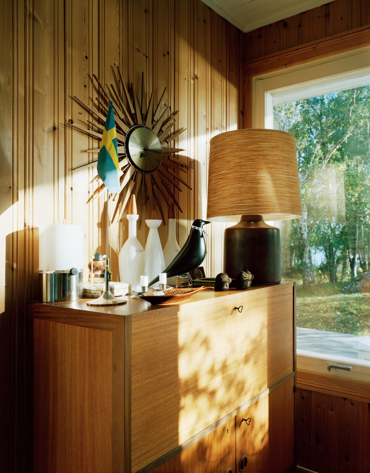 Wooden dresser decorated with a sunburst clock, ceramic lamp, bird figurine, and white vessels against pine-panelled walls.