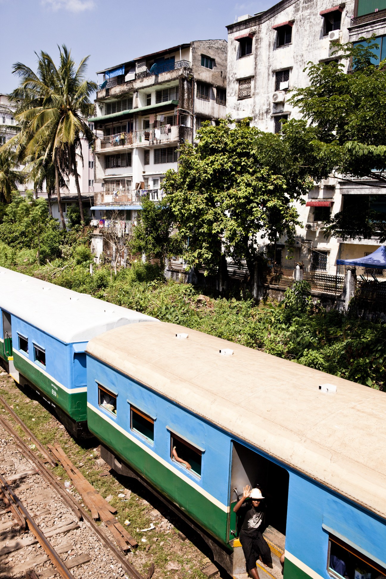 A blue and green train passes colonial buildings overgrown with vegetation in Rangoon.